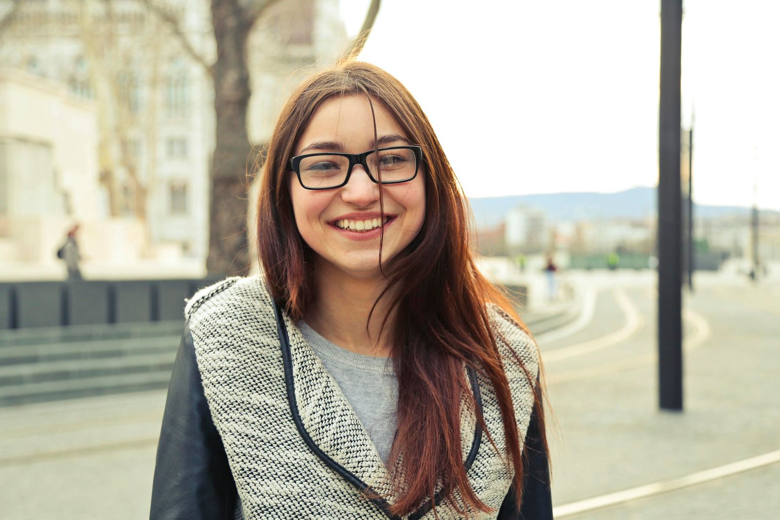 A young woman with glasses smiling outdoors in Budapest street scene, showcasing fashionable style.